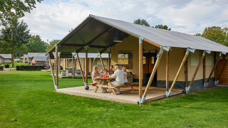 Familia se relaja fuera de una tienda safari Glamping Tent Comfort en un camping verde bajo el cielo azul.