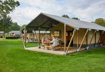 Familia se relaja fuera de una tienda safari Glamping Tent Comfort en un camping verde bajo el cielo azul.