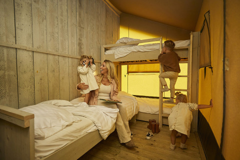 Een gezin geniet samen in een safaritent met stapelbedden, warme gele licht en spelende kinderen