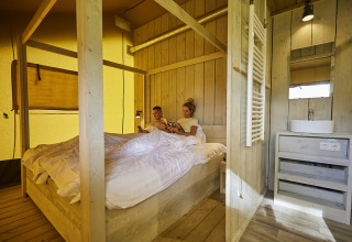 Inside a comfy glamping tent at De Leistert, Netherlands, a couple relaxes together on a cozy wooden bed.
