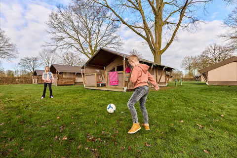 Twee kinderen spelen voetbal voor een Kalahari-safaritent op een grasrijke camping met bomen.