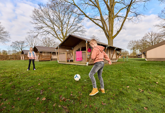Two children playing football outside a Kalahari Safari Tent on a grassy campsite surrounded by trees.
