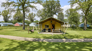Glamping Tent Luxe Safari-Zelt auf einem grünen Campingplatz mit Bäumen und bunten Hütten im Hintergrund.