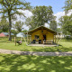 Tienda Glamping Tent Luxe en un camping verde con árboles y cabañas de colores al fondo.