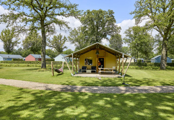 Tienda Glamping Tent Luxe en un camping verde con árboles y cabañas de colores al fondo.