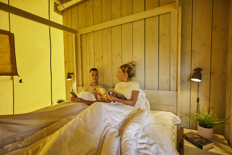Paar liegt entspannt im Bett eines luxuriösen Glampingzeltes mit Holzdekor bei De Leistert, Niederlande.