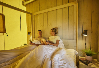 Paar liegt entspannt im Bett eines luxuriösen Glampingzeltes mit Holzdekor bei De Leistert, Niederlande.