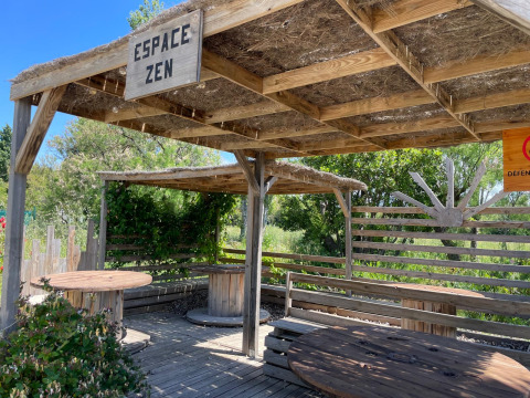 Zona de descanso exterior con bancos de madera y techo de paja en Camping Seasonova Ensoya, Occitania, Francia.