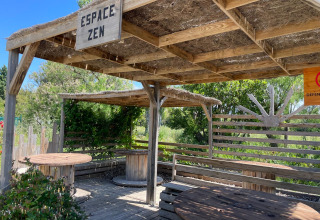 Zona de descanso exterior con bancos de madera y techo de paja en Camping Seasonova Ensoya, Occitania, Francia.
