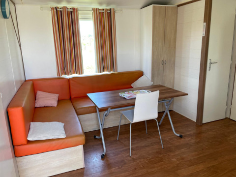 Binnenzicht van een cottage met oranje hoekbank, tafel, stoel en kast bij Camping Seasonova Ensoya, Frankrijk.