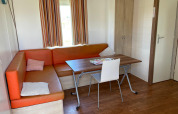 Interior de una cabaña con sofá esquinero naranja, mesa, silla y armario en Camping Seasonova Ensoya, Francia.