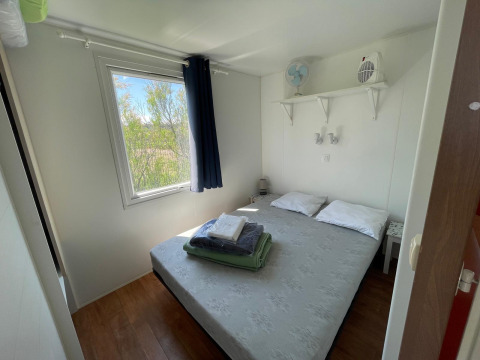 Lichte slaapkamer met tweepersoonsbed, raam, plankjes en airconditioning in een huisje bij Camping Seasonova Ensoya, Frankrijk.