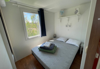 Lichte slaapkamer met tweepersoonsbed, raam, plankjes en airconditioning in een huisje bij Camping Seasonova Ensoya, Frankrijk.