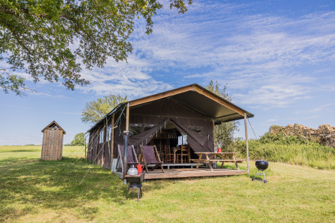 Safari-Zelt Glampinglodge de luxe mit privatem Bad auf einer grünen Wiese unter blauem Himmel.