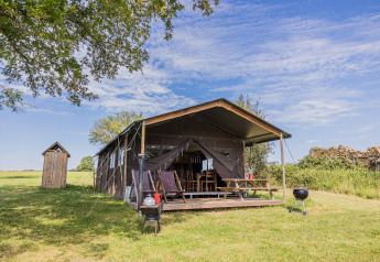 Safari-Zelt Glampinglodge de luxe mit privatem Bad auf einer grünen Wiese unter blauem Himmel.