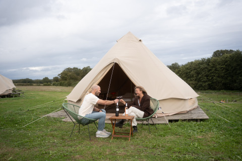 Two people sit in front of a Bell tent teepee, toasting with wine outdoors in a scenic grassy campsite.