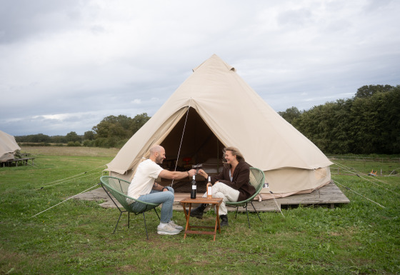 Dos personas brindan con vino frente a una tienda tipi Bell en un campamento verde y natural al aire libre.