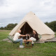 Two people sit in front of a Bell tent teepee, toasting with wine outdoors in a scenic grassy campsite.