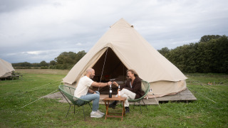 Two people sit in front of a Bell tent teepee, toasting with wine outdoors in a scenic grassy campsite.
