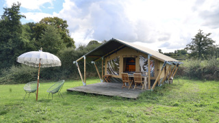 Tienda safari con instalaciones sanitarias privadas en un campo verde, con muebles y sombrilla afuera.