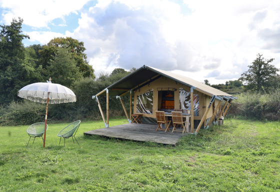 Tenda safari con servizi igienici privati su prato verde, veranda in legno e arredi all'aperto.