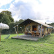 Tienda safari con instalaciones sanitarias privadas en un campo verde, con muebles y sombrilla afuera.