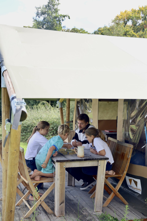 Family playing a board game at a wooden table outside a safari tent with private facilities in France.