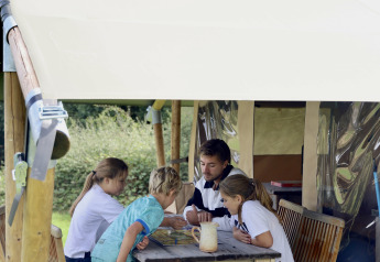 Family playing a board game at a wooden table outside a safari tent with private facilities in France.