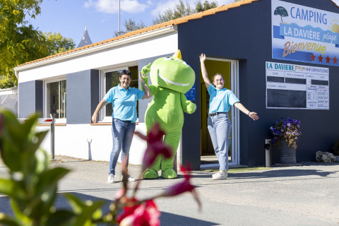 Two staff members and a green mascot welcome guests outside Flower Camping La Davière Plage in Pays de la Loire, France.