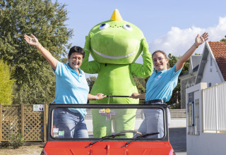Drie personen, waaronder een groene mascotte, staan in een rode auto bij Flower Camping La Davière Plage.