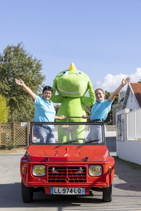 Drie personen, waaronder een groene mascotte, staan in een rode auto bij Flower Camping La Davière Plage.