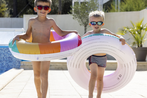 Dos niños con gafas de sol y flotadores caminan junto a la piscina en Flower Camping La Davière Plage, Francia.