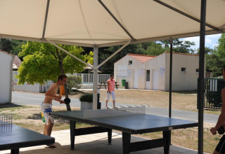 To personer spiller bordtennis udendørs under et halvtag på Flower Camping La Davière Plage, Frankrig.
