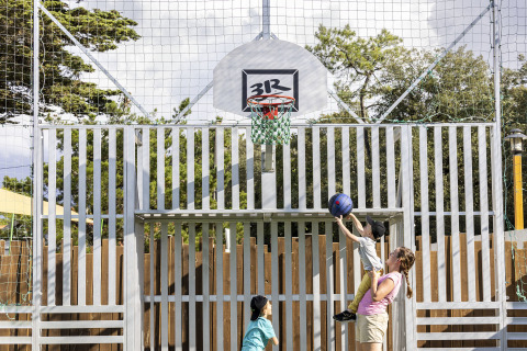 Un adulto aiuta un bambino a tirare a canestro su un campo da basket all’aperto, osservato da un altro bimbo.