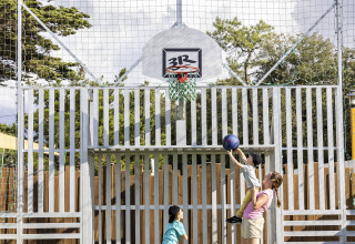 An adult helps a child shoot a basketball at an outdoor court while another child watches nearby.