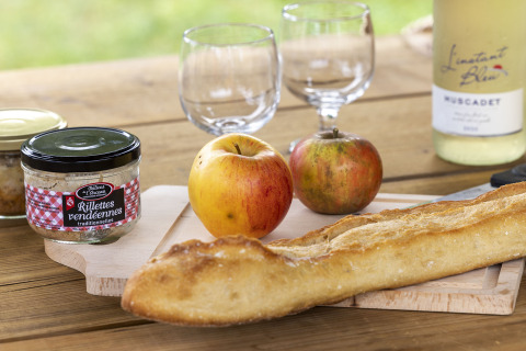 Mesa rústica con baguette, manzanas, rilletes, vino y copas en Flower Camping La Davière Plage, Francia.