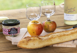 Mesa rústica con baguette, manzanas, rilletes, vino y copas en Flower Camping La Davière Plage, Francia.