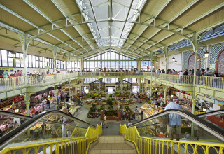 Spacious two-level indoor market at Flower Camping La Davière Plage, with food and flower stalls, viewed from an escalator.