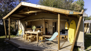Tenda safari Hut Cotton Confort con veranda in legno, tavolo, sedie e tavola da surf all’esterno.