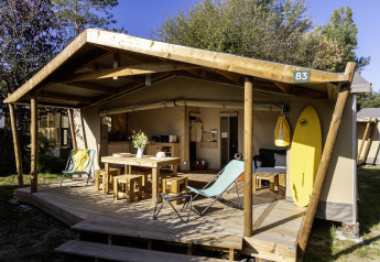 Tenda safari Hut Cotton Confort con veranda in legno, tavolo, sedie e tavola da surf all’esterno.