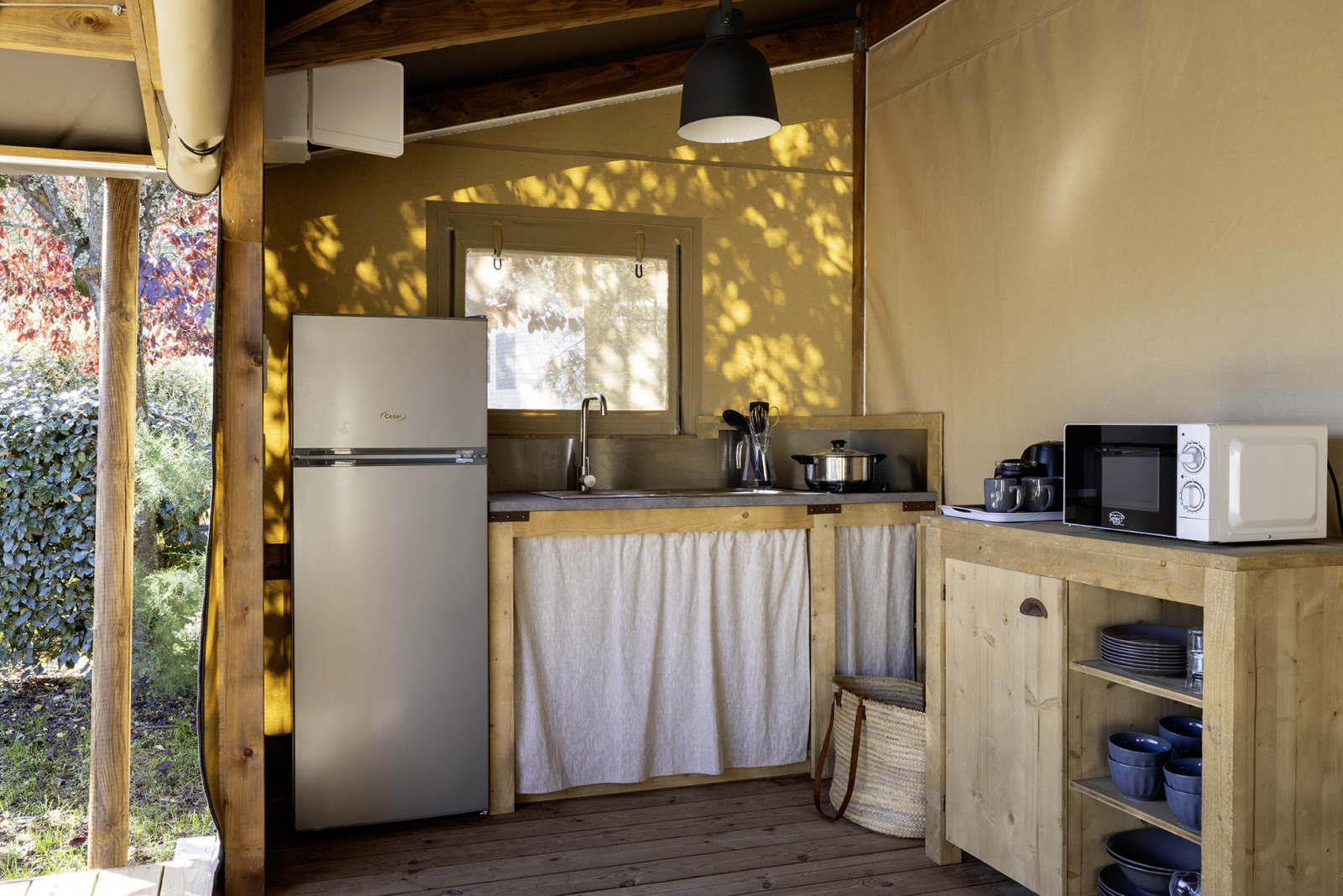 Keukengedeelte in een safaritent met houten meubels, koelkast, magnetron en servies in natuurlijk licht.