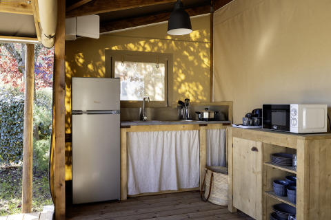 Küche in einem Safari-Zelt mit Holzmöbeln, Kühlschrank, Mikrowelle und Geschirr im Sonnenlicht.