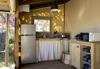 Angolo cucina in una tenda safari con mobili in legno, frigorifero, microonde e stoviglie alla luce naturale.