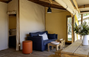 Woonruimte in safari tent Hut Cotton Confort met zetel, houten meubilair en bloemen op de tafel.