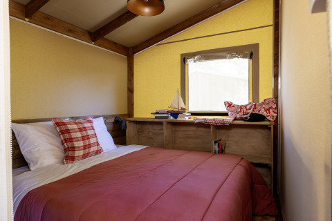 Schlafzimmer in einem Safarizelt mit Holzbalken, Doppelbett, roter Bettdecke und Fenster in La Davière Plage.