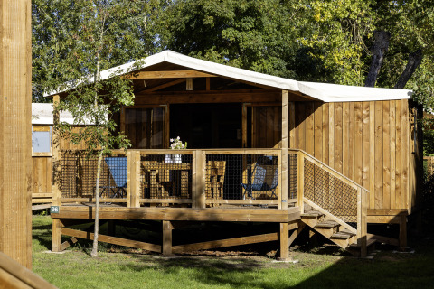 Safari-Zelt CosyFlower Confort mit Holzveranda, Sitzgelegenheiten und von Bäumen umgeben.