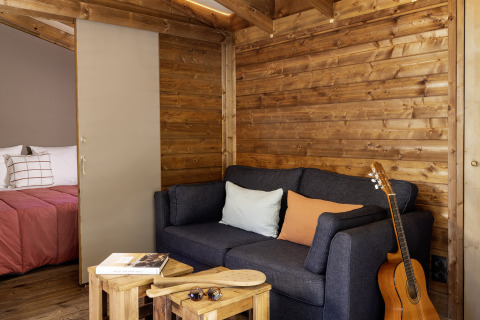 Gemütlicher Wohnbereich im Safarizelt mit Holzwänden, dunkelgrauem Sofa, Gitarre und Blick ins Schlafzimmer.