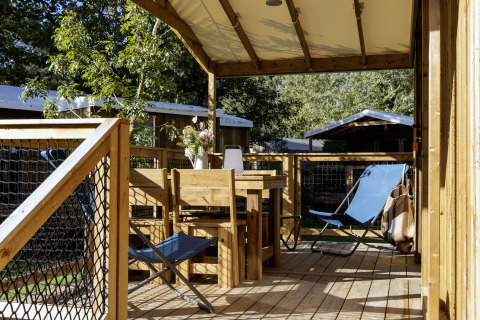 Überdachte Terrasse mit Holzmöbeln und blauem Liegestuhl am CosyFlower Confort Safarizelt in Frankreich.