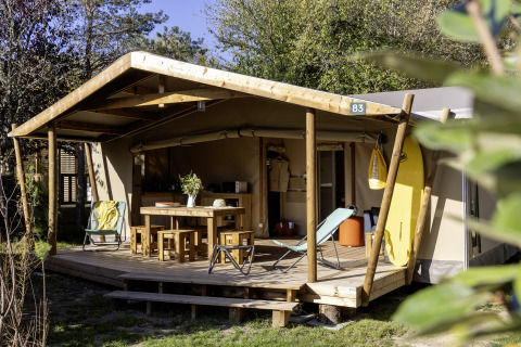 Safari-Zelt Hut Cotton Confort mit Terrasse, Holzmöbeln und Surfbrett in einer grünen, sonnigen Umgebung.