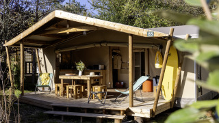 Safari tent Hut Cotton Confort met houten terras, tuinmeubilair en surfplank in een natuurlijke omgeving.